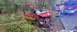 Na zdjęciu widać przód rozbitego pojazdu biorącego udział w zdarzeniu. Obok samochodu stoją dwaj strażacy a w tle widać samochód pożarniczy. Zdarzenie miało miejsce na drodze przebiegającej przez las