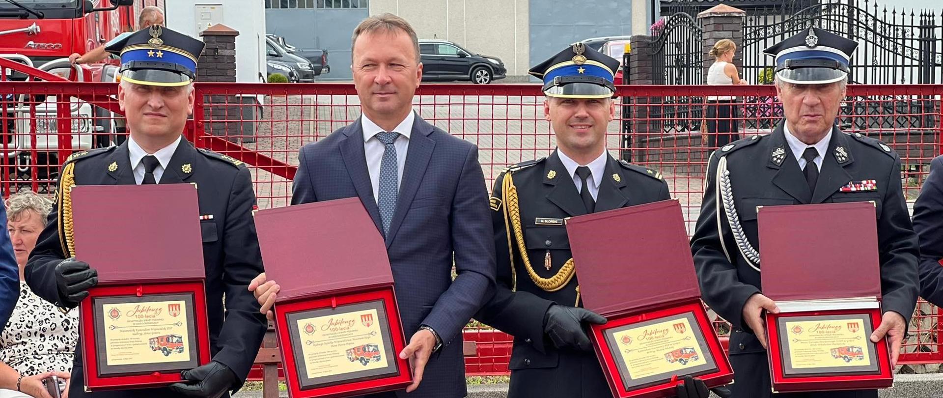 Trzej strażacy oraz jeden mężczyzna w garniturze, prezentujący pamiątkowe tablice