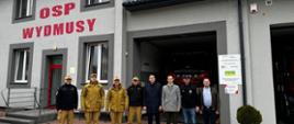 Strażacy i władze samorządu terytorialnego z Myszyńca i Gminy Rozogi stoją w rzędzie przed remizą OSP Wydmusy podczas inauguracji Posterunku Czasowego JRG PSP w Ostrołęce na bazie remizy OSP Wydmusy