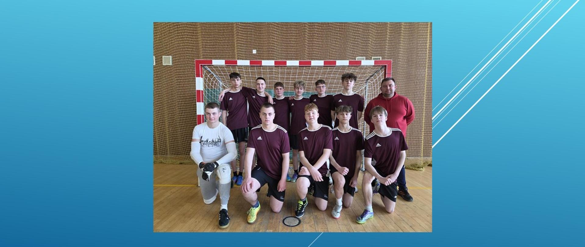 Zawodnicy i trener pozują do zdjęcia na tle bramki w hali sportowej.