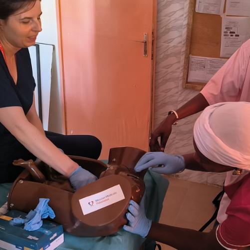 Trzy kobiety w strojach pracowników medycznych. Dwie wykonują ćwiczenia na fantomie, trzecia się przygląda