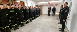 Na zdjęciu w sali konferencyjnej Komendy Powiatowej Państwowej Straży Pożarnej widoczni strażacy Państwowej Straży Pożarnej i druhowie kończący szkolenie ubrani w ubrania koszarowe w czarnym kolorze podczas uroczystego apelu kończącego szkolenie.