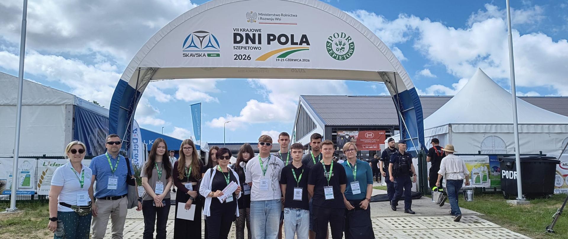 Na zdjęciu widać grupę kilkunastu osób stojących przed głównym wejściem na targi lub wydarzenie branżowe. Nad uczestnikami znajduje się duży biały łuk z napisem „VII Krajowe Dni Pola Szepietowo 2026” oraz logotypami organizatorów i partnerów wydarzenia. Wszyscy mają na szyjach identyfikatory, co sugeruje udział w wycieczce edukacyjnej, szkoleniu lub konferencji.