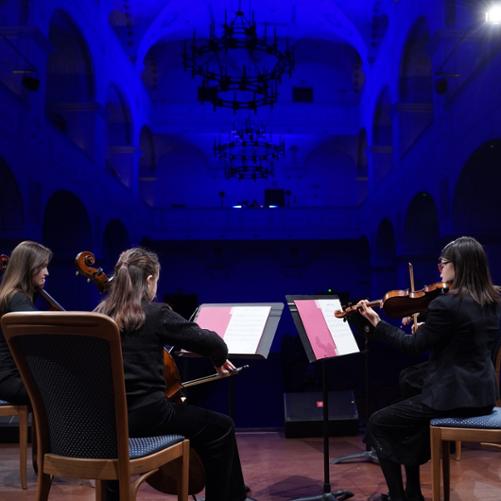 zdjęcie przedstawia trzy uczennice z kwartetu smyczkowego podczas wykonania utworu na scenie sali koncertowej, ujęcie od tyłu z widokiem na przestrzeń sali koncertowej