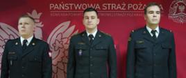 Świetlica Komendy Powiatowej PSP w Rawiczu. Na tle ścianki promocyjnej stoją w szeregu trzej awansowani strażacy. Mają na sobie mundury wyjściowe. 
