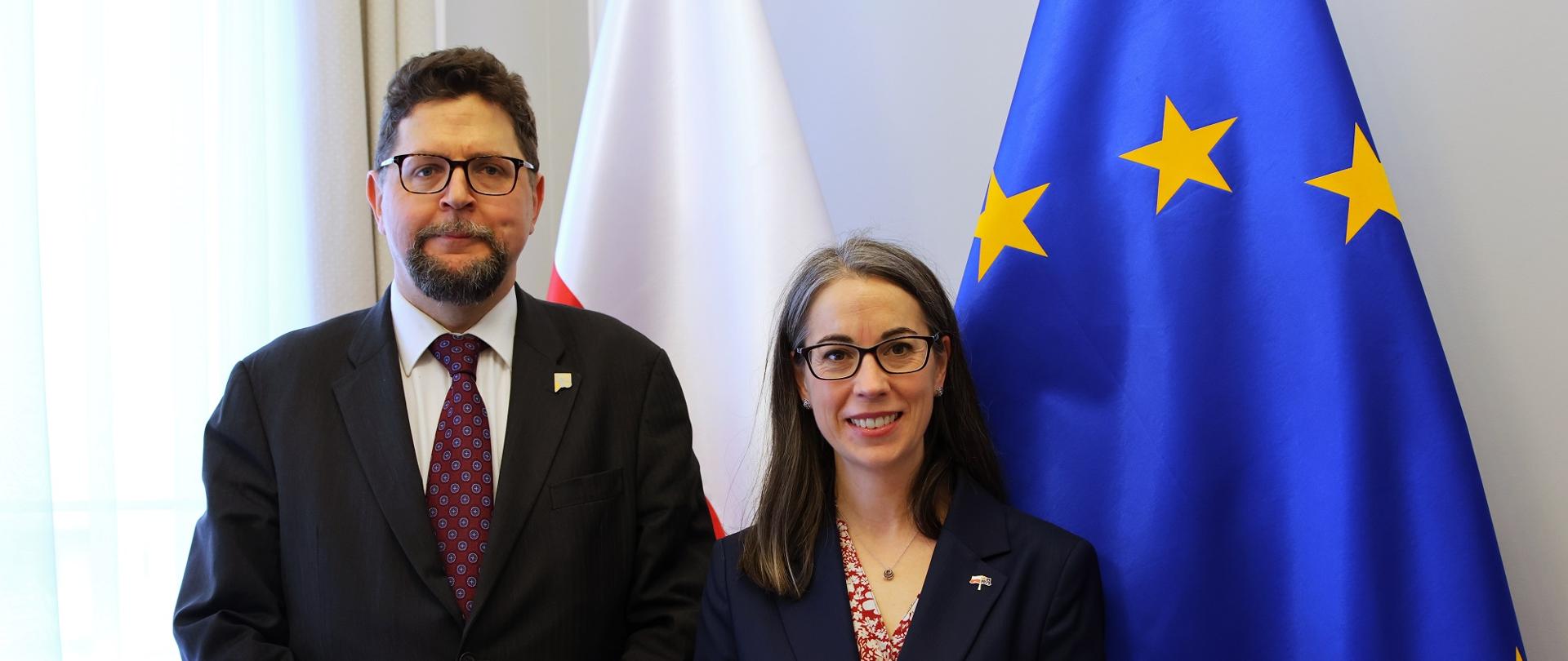 Pod ścianą stoi wiceminister Szeptycki i kobieta w czarnej marynarce, za nimi flagi Polski i UE.