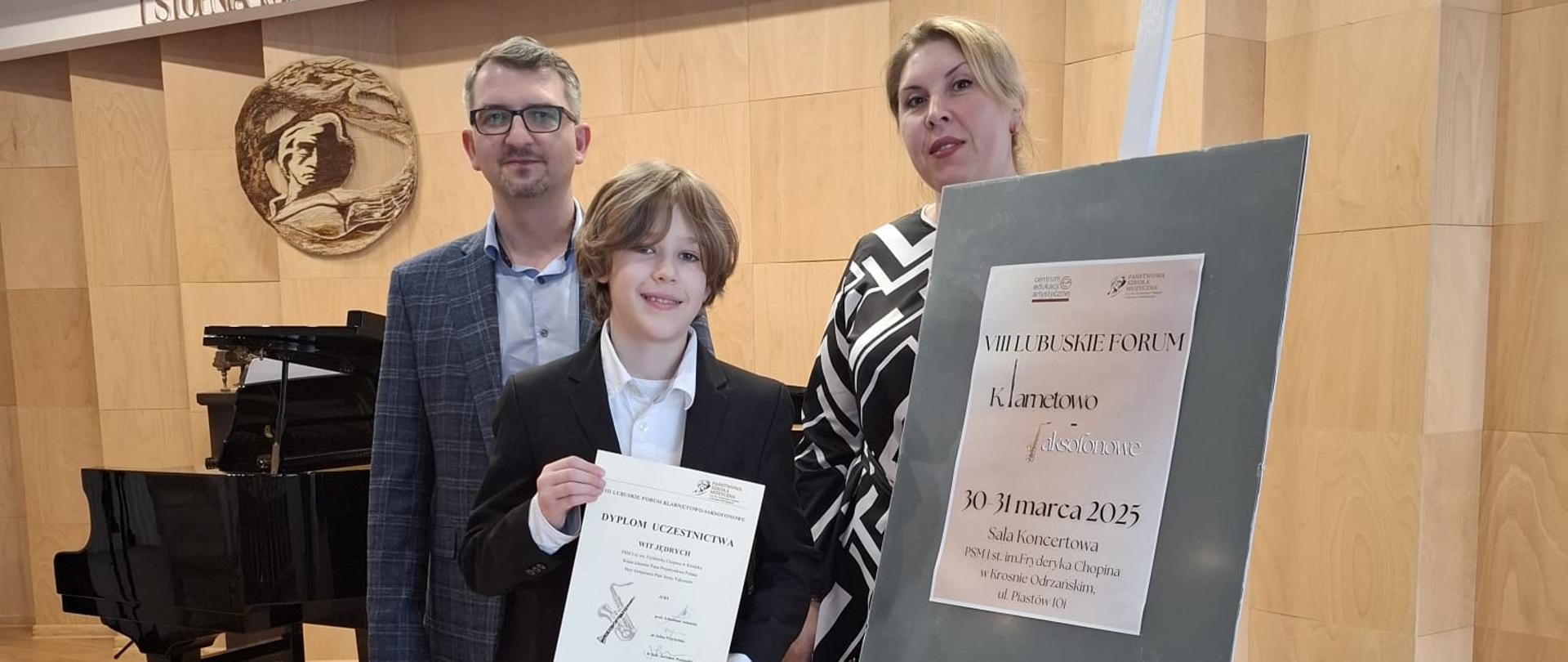Nauczyciel klarnetu Przemysław Polak oraz nauczyciel fortepianu Iryna Yakymets wraz z uczniem Witem Jędrych pozują do zdjęcia na scenie sali koncertowej w Krośnie Odrzańskim