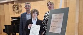 Nauczyciel klarnetu Przemysław Polak oraz nauczyciel fortepianu Iryna Yakymets wraz z uczniem Witem Jędrych pozują do zdjęcia na scenie sali koncertowej w Krośnie Odrzańskim