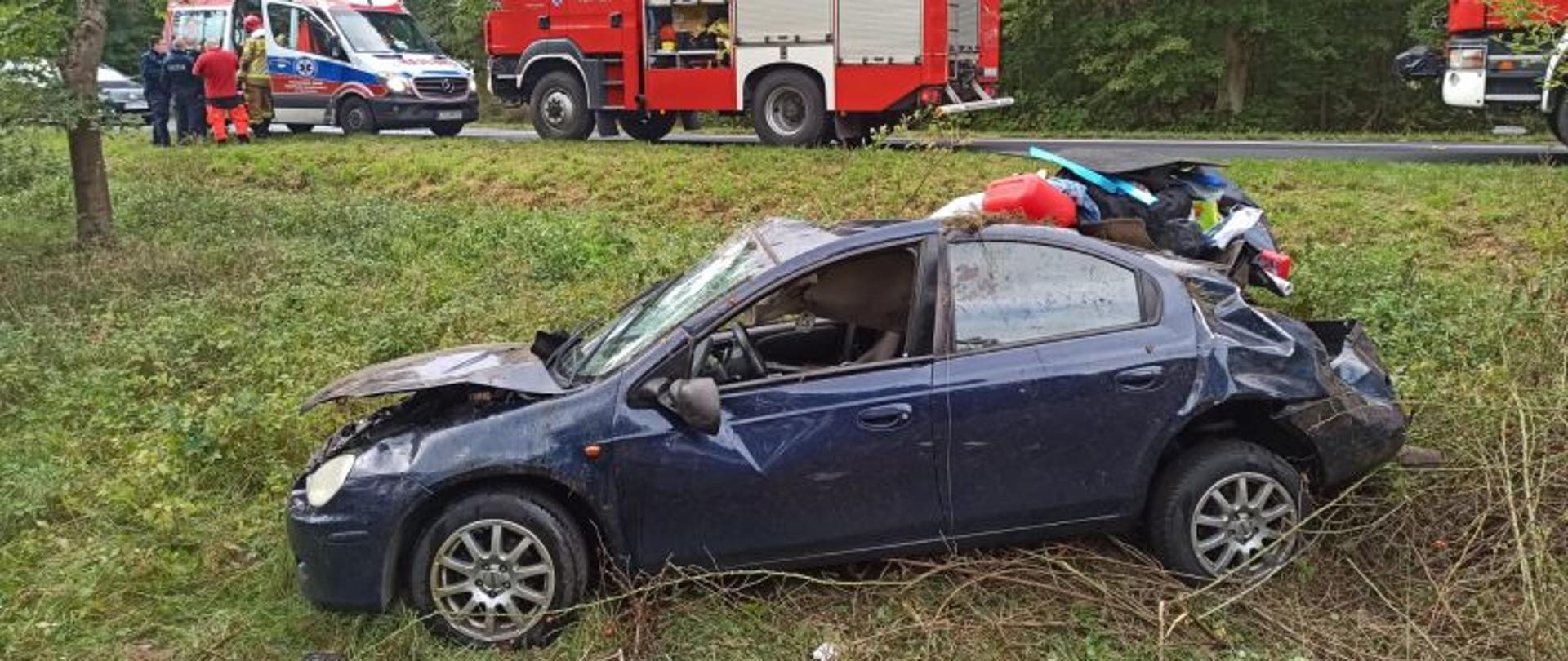 Na pierwszym planie auto poza jezdnią, stoi wzdłuż drogi. Auto po dachowaniu, zniszczone z każdej strony. Auto koloru ciemno niebieskiego. Na drugim planie, na drodze widać stojące dwa pojazdy pożarnicze oraz karetkę pogotowia. Przy karetce stoją policjanci, ratownik medyczny i strażak