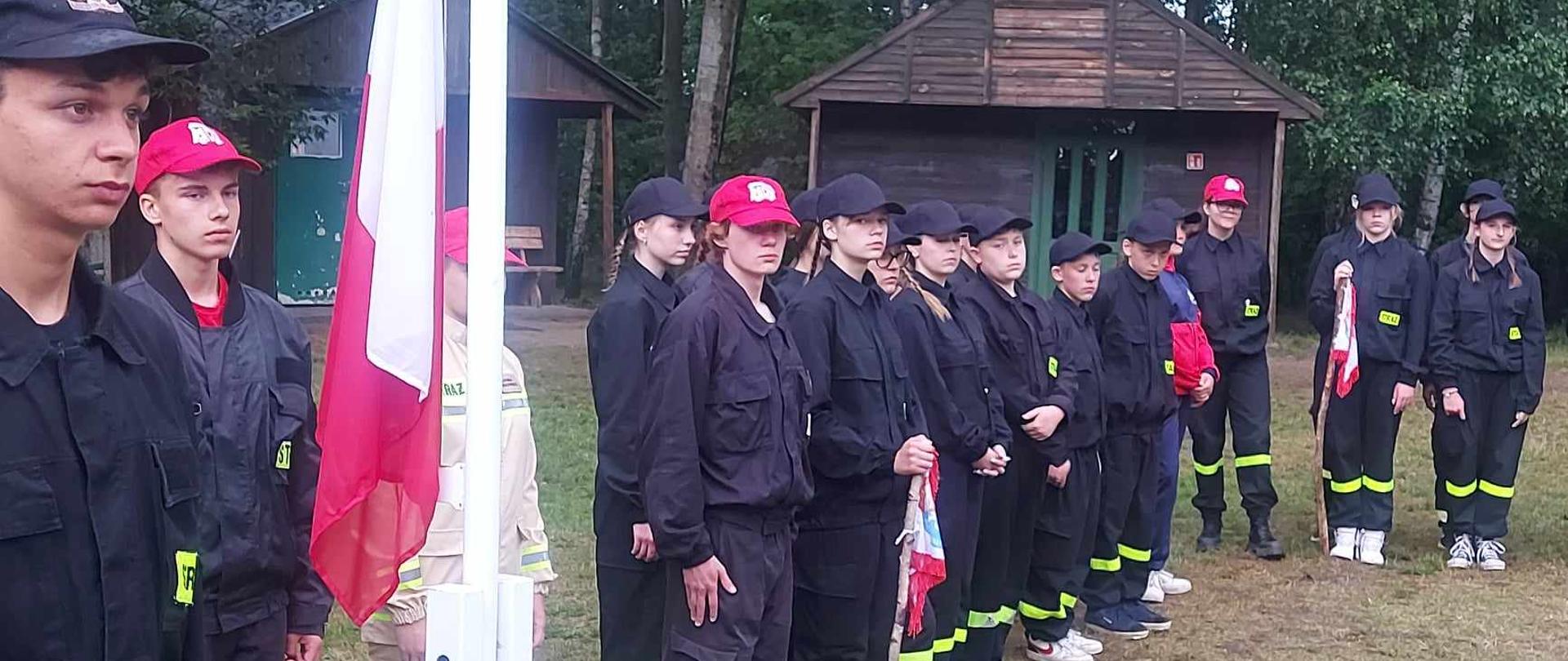 Zdjęcie przedstawia strażaków oraz dzieci i młodzież na wakacyjnym obozie strażackim.
W tle ośrodek wypoczynkowy.
