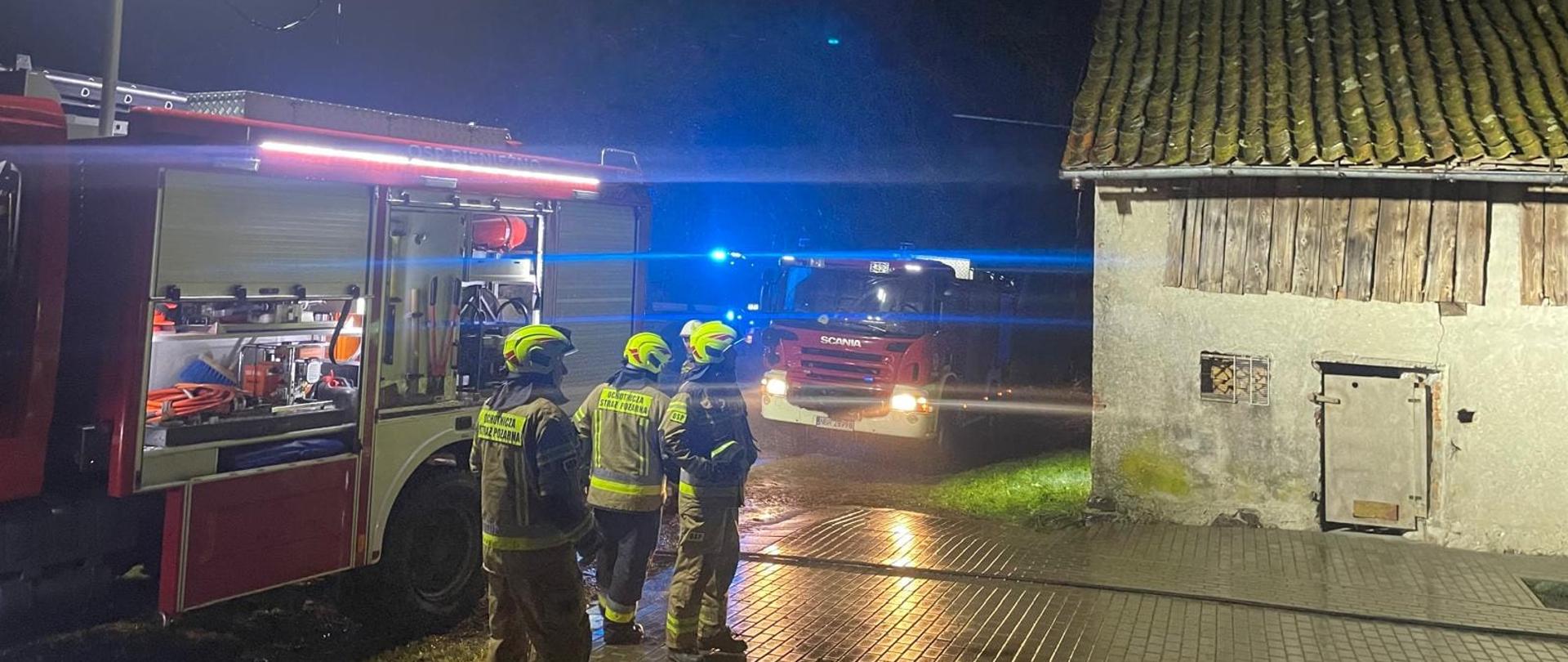 Zdjęcie zrobione nocą. Z lewej strony stoją samochody strażackie oświetlają miejsce zdarzenia. Z prawej strony budynek.
