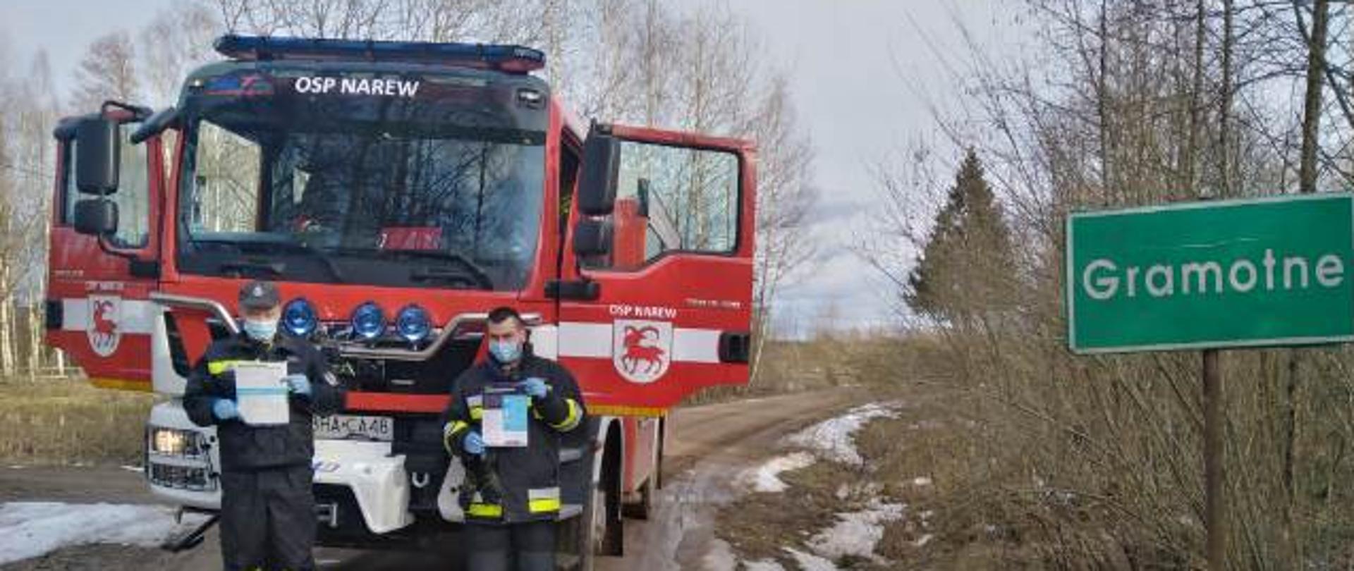 Widoczny pojazd gaśniczy uczestniczący w kolportażu ulotek na terenie gminy Narew, droga żwirowa, ratownicy z ulotkami na tle samochodu gaśniczego i tablicy z miejscowością Gramotne