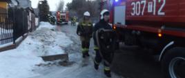 Zdjęcie przedstawia strażaków, którzy ze zwiniętymi wężami idą w kierunku samochodu strażackiego. W tle widać drugi samochód straży pożarnej i grupę strażaków.
