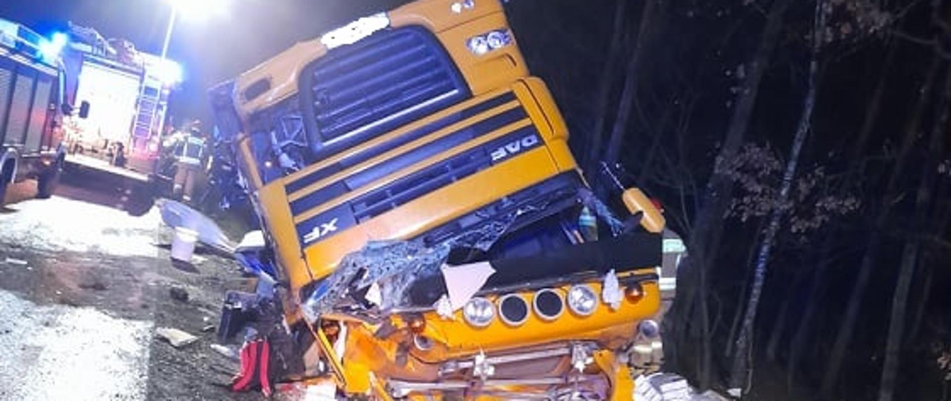 Na zdjęciu widoczny żółty ciągnik siodłowy leżący do góry kołami oraz wysypany materiał. W tle samochody straży pożarnej.
