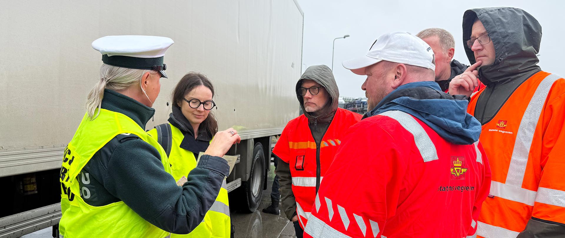 Inspektor ITD mówi do norweskich gości. Norwegowie słuchają i patrzą na to, co pokazuje polski inspektor. Za nimi pojazd ciężarowy z białą naczepą.