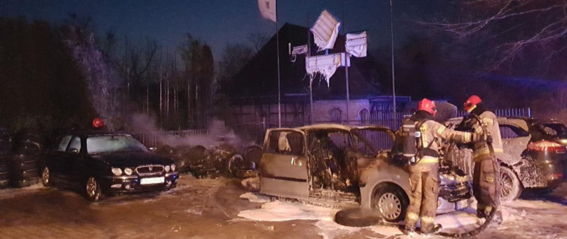 Zdjęcie przedstawia pojazdy znajdujące się na placu przy warsztatowym wykonane nocą. Od lewej strony zdjęcia widoczny czarny pojazd marki Rover, po prawej stronie dwa spalone pojazdy marki Peugeot i Ford. Przed pojazdami dwaj strażacy w ubraniach specjalnych koloru piaskowego w aparatach powietrznych gaszą z węża pożarniczego wnętrze komory silnikowej