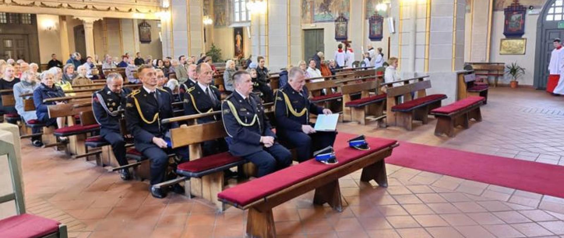Zdjęcie wykonane wewnątrz kościoła. Widok skierowany jest od strony ołtarza na wnętrze. Widać ławki, wiernych. W pierwszych rzędach strażacy w galowych mundurach.