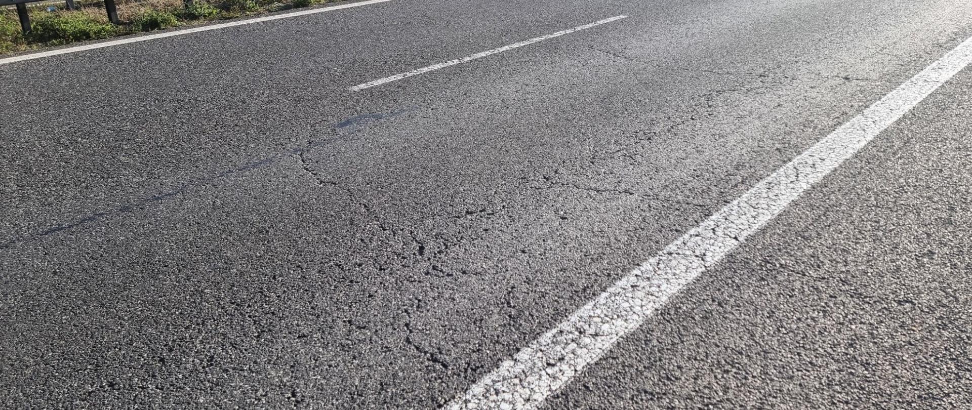 Fragment jezdni autostrady z widocznymi pęknięciami nawierzchni. Obok drogi znajduje się bariera ochronna, a na sąsiedniej jezdni przejeżdżają samochody. W tle widoczny jest nasyp porośnięty trawą.