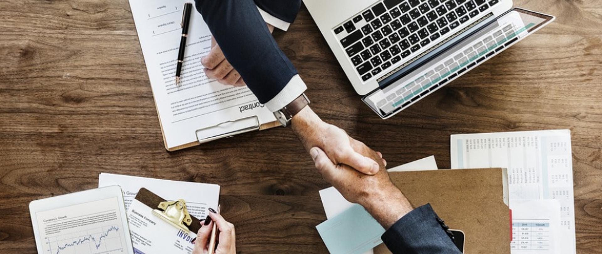 Dwaj mężczyźni ściskają sobie dłonie, obok otwartego laptopa i rozłożonych dokumentów.