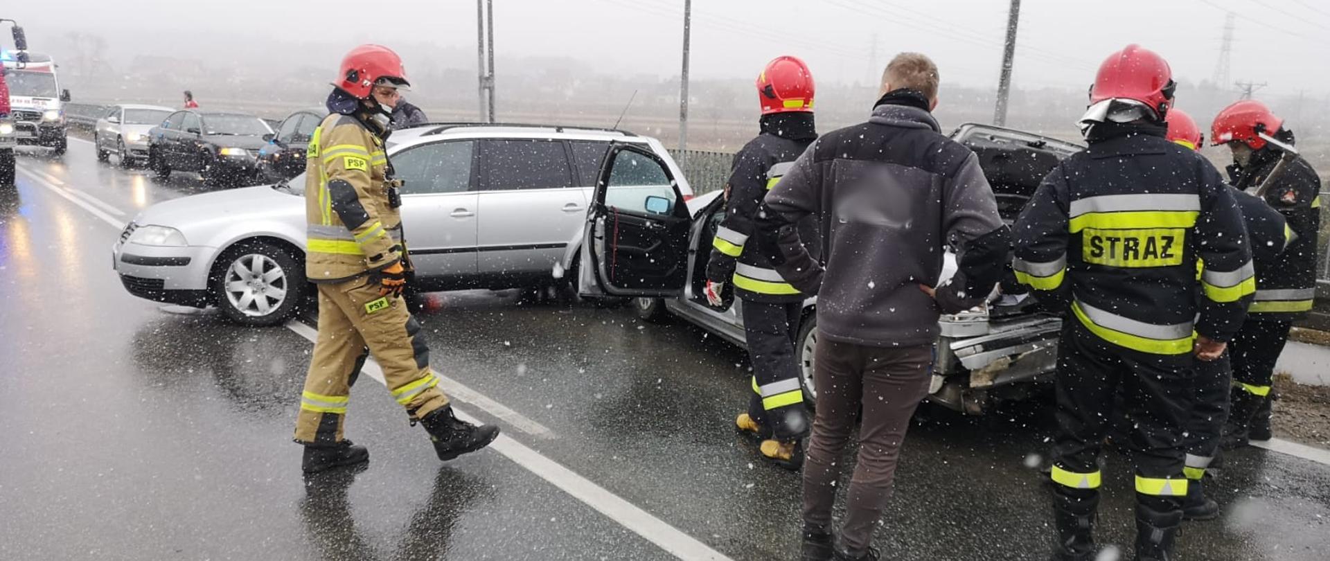 Dwa samochody osobowe stoją na drodze. Jeden stoi w poprzek drogi. Obok samochodów strażacy prowadzący działania i jedna osoba postronna.