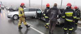Dwa samochody osobowe stoją na drodze. Jeden stoi w poprzek drogi. Obok samochodów strażacy prowadzący działania i jedna osoba postronna.