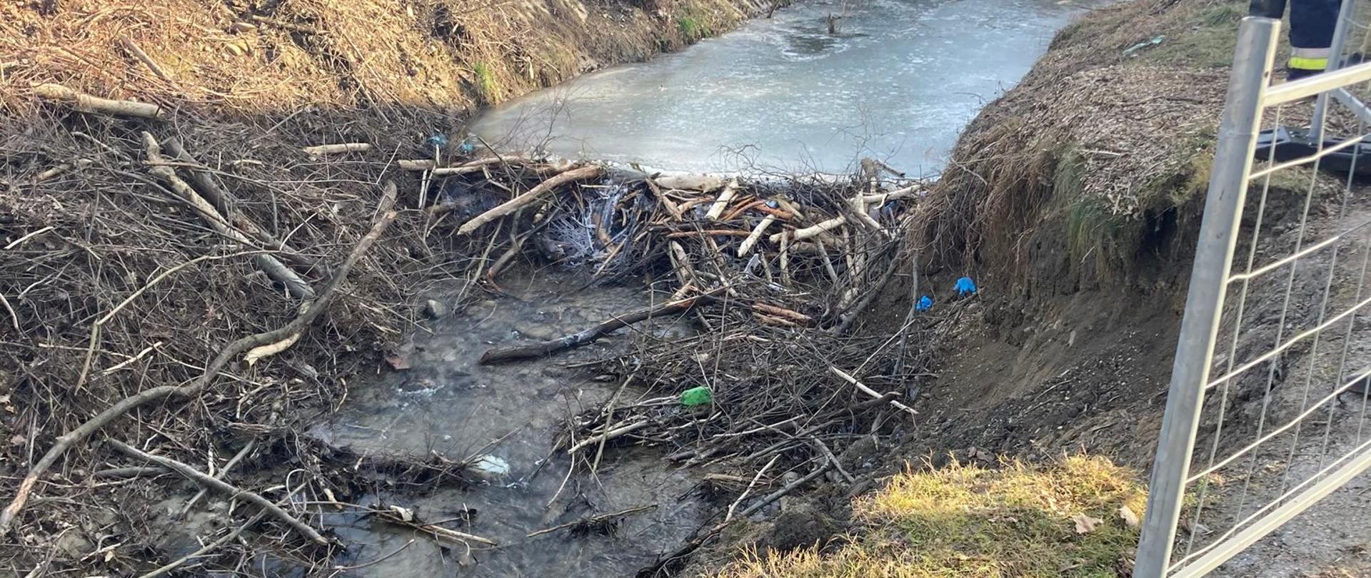 Potok częściowo skuty lodem oraz fragment barierki drogowej