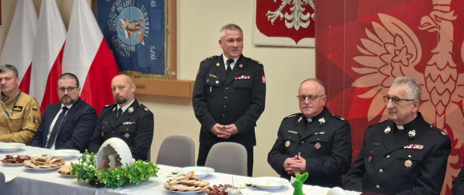 Przy stołach na których znajdują się dekoracje świąteczne i talerze, siedzą strażacy i zaproszenie goście. Strażacy ubrani są w mundury galowe zaproszeni goście odświętnie w garniturach. Komendant w postawie stojącej przemawia. W tle logo Państwowej Straży Pożarnej oraz godło Polski i flagi