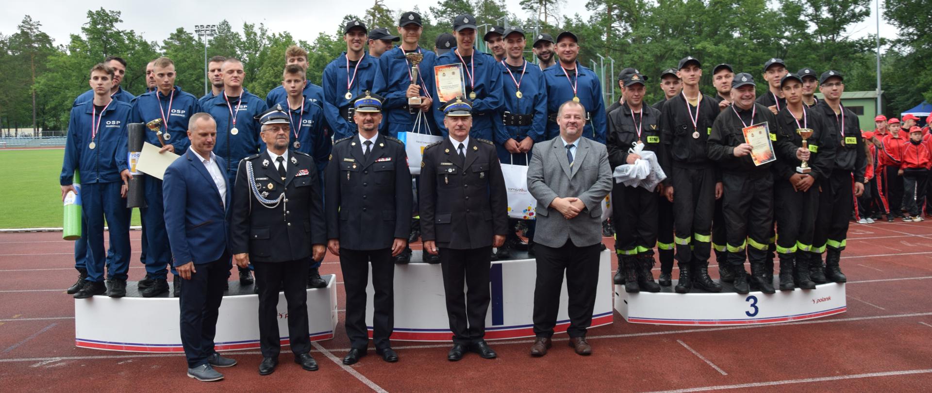 trzy drużyny strażaków ochotników stojące na poszczególnych stopniach podium, dwie w niebieskich jednokolorowych strojach trzeci drużyna w mundurach koszarowych, przed nimi stoi 3 strażaków w umundurowaniu wyjściowym i dwóch mężczyzn w garniturach, w tle drzewa