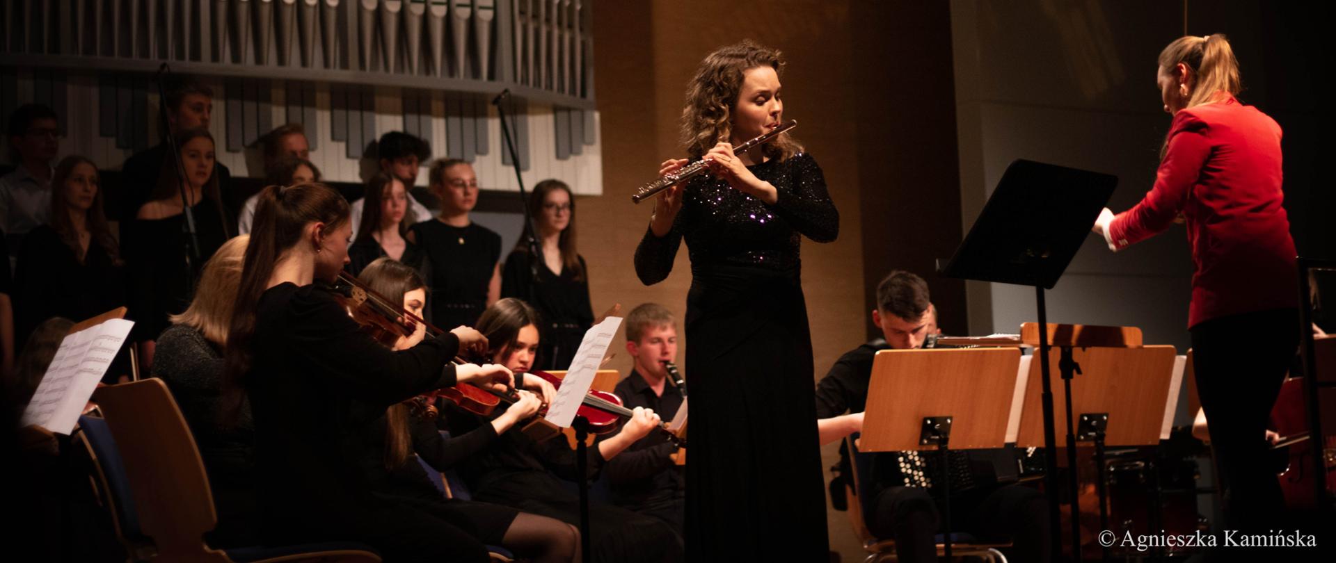 Zdjęcie z koncertu, przedstawiające realizującą solo flecistkę, obok Martyna Lasota - prowadząca orkiestrę i chór. 