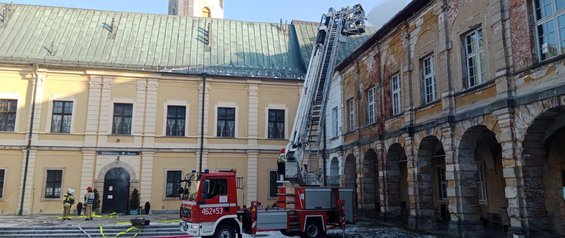 Widok na zamek i plac zamkowy. Na placu stoi podnośnik z wysuniętym koszem, w którym znajduje się strażak prowadzący działania gaśnicze. Po lewej stronie przed drzwiami do budynku stoi trzech strażaków w umundurowaniu specjalnym.