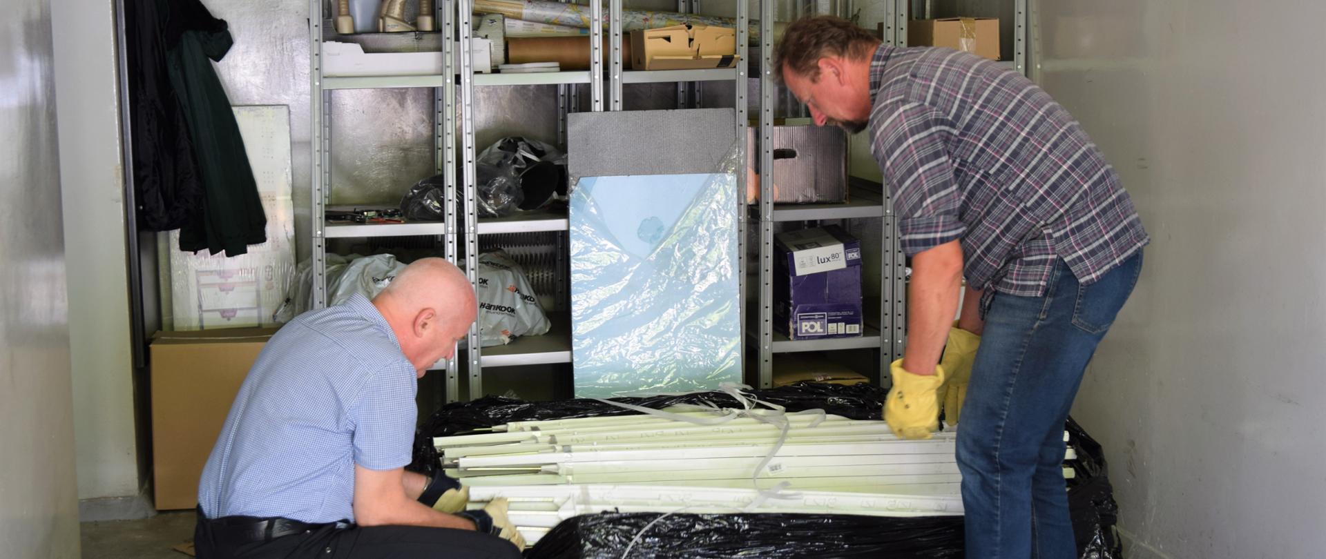 Dwóch mężczyzn, stojących po prawej i lewej stronie, nachylających się nad plastikowymi elementami, wystającymi z czarnej folii. W tle szafka zbudowana z metalowego stelażu, a na pólkach stoją m.in. kartony.