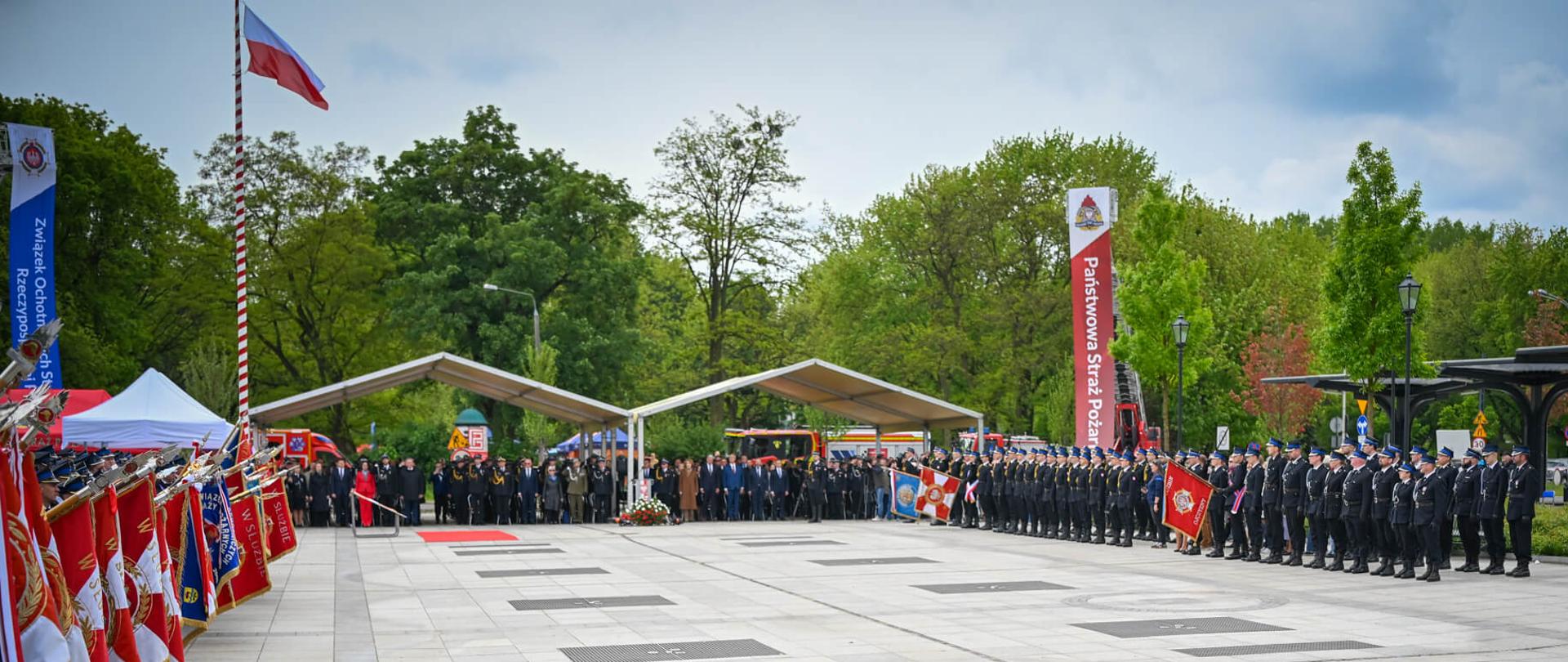 Perspektywiczne ujęcie placu podczas oficjalnej uroczystości. Po lewej stronie widać liczne poczty sztandarowe, po prawej rzędy strażaków, a w głębi trybunę z gośćmi honorowymi pod zadaszeniem.