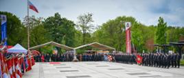 Perspektywiczne ujęcie placu podczas oficjalnej uroczystości. Po lewej stronie widać liczne poczty sztandarowe, po prawej rzędy strażaków, a w głębi trybunę z gośćmi honorowymi pod zadaszeniem.