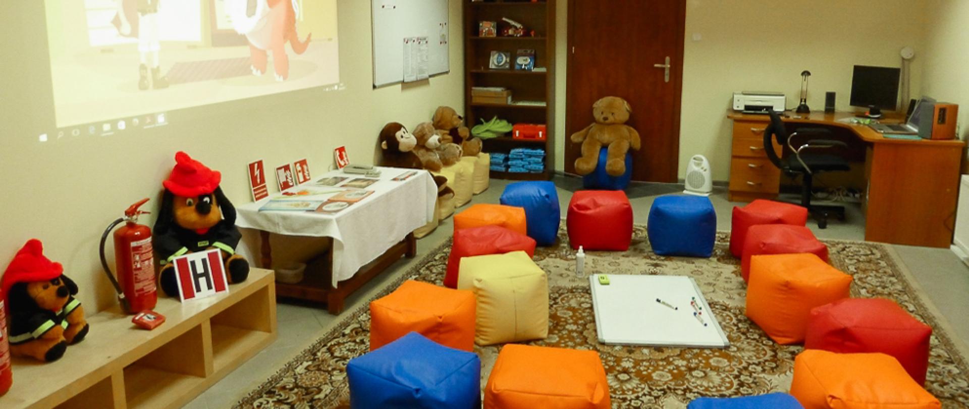 Sala edukacyjna do zajęć z dziećmi. Po ścianą zabawki pluszowe. Na podłodze kolorowe pufy. Na ścianie wyświetlany jest film edukacyjny dla dzieci.
