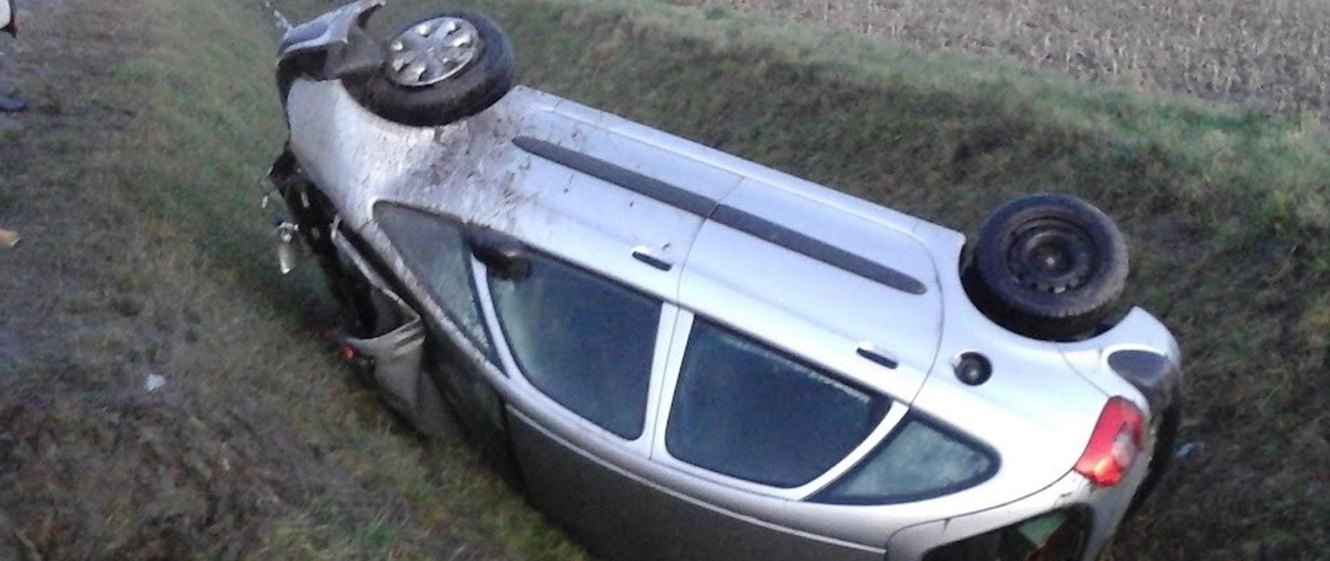 Uszkodzony samochód osobowy leży na dachu w przydrożnym rowie.