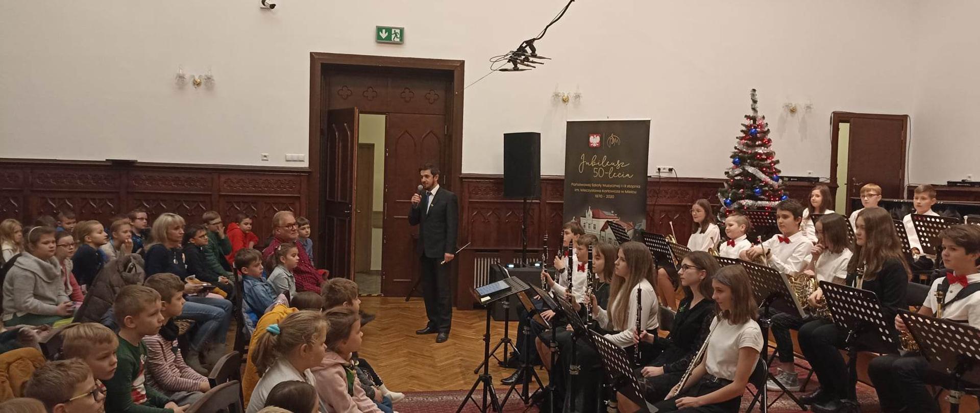 zdjęcie z audycji dla najmłodszych uczniów szkół podstawowych przedstawiające orkiestrę dętą PSM I stopnia naszej szkoły z dyrygentem Piotrem Rysiewiczem na przedzie oraz uczestników audycji z lewej strony zdjęcia