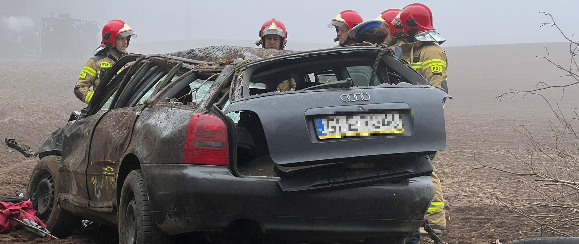 Zdjęcie przedstawia rozbity samochód osobowy Audi A4 kombi (po dachowaniu) stojący na polu oraz strażaków prowadzących czynności służbowe.