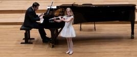 Dziewczynka stojąc na środku estrady sali koncertowej PSM gra na flecie, za nią na fortepianie gra mężczyzna.
