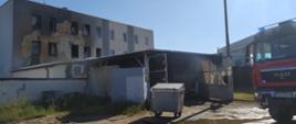 Pożar kompleksu budynków. Widok na parterowy budynek magazynowy, w którym powstał pożar. Przed budynkiem jest wiata, od której otwarta jest brama. Przed wiatą stoi metalowy kontener na odpady. Z prawej strony widoczny przód wozu strażackiego. W tle hotel ze zniszczona elewacją zewnętrzną.