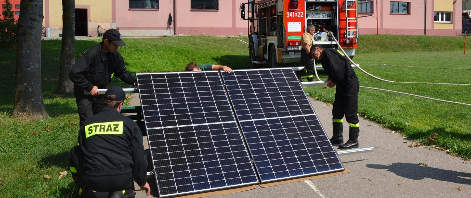 Zdjęcie obrazuje strażaków oglądających panele fotowoltaiczne w głębi samochód strażacki i budynek JRG nr 1.