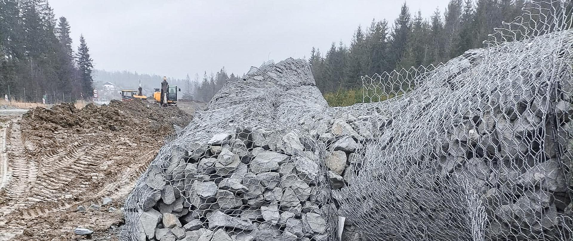 Na zdjęciu widzimy fragment zdegradowanej ściany gabionowej. Konstrukcja, niegdyś stabilizująca nasyp, teraz jawi się jako grupa rozproszonych metalowych koszy, wypełnionych nieregularnie ułożonymi kamieniami. Kilka z górnych koszy zostało wyraźnie uszkodzonych, co skutkuje przewróceniem się części gabionów. W tle migruje klimat miejsca budowy – sprzęt roboczy delikatnie kontrastuje z chaotycznie rozmieszczonymi elementami struktury, a przestrzeń wokół nadaje całości surowy, nieuporządkowany charakter. Fotografię można odczytać jako wizualną dokumentację degradacji konstrukcji wynikającej z ingerencji sił zewnętrznych.
