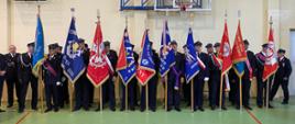 Druhowie OSP z jednostek OSP powiatu ciechanowskiego ze sztandarami przed rozpoczęciem uroczystego apelu na hali sportowej w Gąsocinie. Wszyscy ubrani w mundury. 