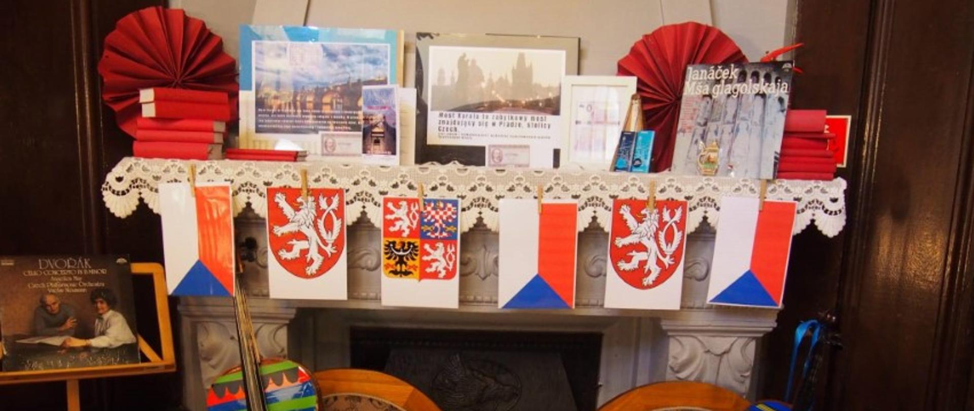 Zdjęcie kolorowe. Kompozycja tematyczna poświęcona Czechom ustawiona przy kominku. Na białej, koronkowej serwecie leżącej na gzymsie stoją książki w czerwonych oprawach, czerwone papierowe wachlarze, fotografie czeskich zabytków z opisami, drobne pamiątki oraz płyta winylowa Janáčka. Do krawędzi serwety przypięte klamerkami papierowe flagi Czech oraz herby z lwem i orłem. Poniżej, na dwóch tapicerowanych fotelach o brązowym wzorze, oparte dwa instrumenty smyczkowe (wiolonczele) pomalowane w jaskrawe, poziome pasy. Po lewej stronie nutnik z okładką albumu Dvořáka. W tle ciemne, drewniane drzwi i ozdobna obudowa kominka.