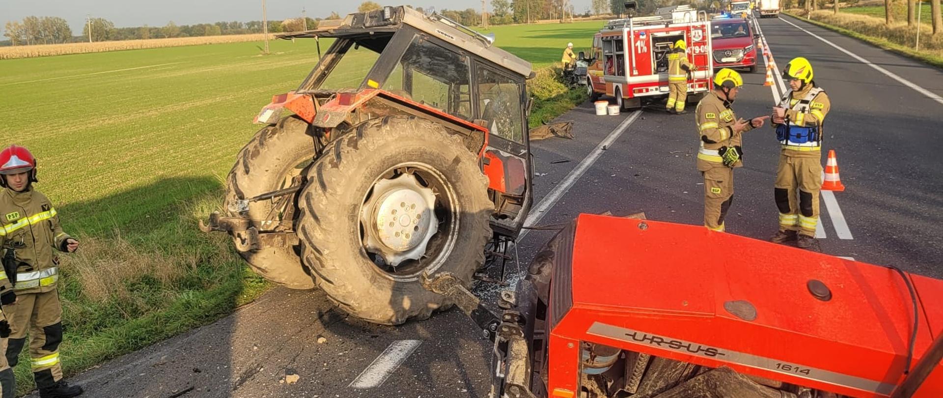 Na zdjęciu widoczna jest scena poważnego wypadku drogowego z udziałem ciągnika rolniczego marki Ursus, który uległ rozczepieniu – jego tylna część stoi na poboczu, natomiast przednia leży na środku drogi. Na miejscu pracują strażacy ubrani w pełne umundurowanie ochronne i hełmy, zabezpieczając miejsce zdarzenia. W tle widoczny jest średni samochód ratowniczo-gaśniczy, radiowóz oraz karetka pogotowia. Część drogi jest zablokowana, ustawiono pachołki i taśmy ostrzegawcze. Na jezdni widać rozlany olej oraz rozsypane elementy konstrukcyjne pojazdu. Akcja odbywa się w ciągu dnia przy dobrej pogodzie, w otoczeniu pól i drzew przydrożnych.