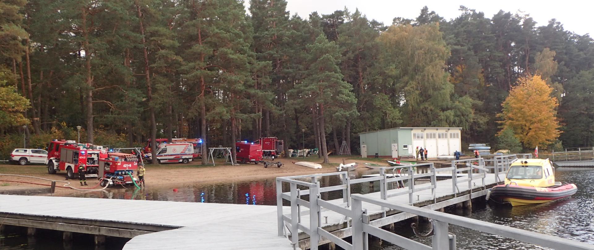 Widok na pomost na jeziorze a w tle pojazdy strażackie oraz pogotowia ratunkowego stoją nad brzegiem jeziora.