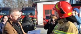 Senator Jarosław Rusiecki wręcza dowódcy JRG Nr 1 w Ostrowcu Św. akt przekazania samochodu. W tle fotoreporterzy i zaparkowane na placu samochody strażackie.