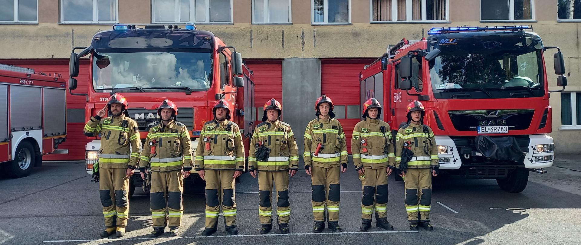 Zdjęcie przedstawia strażaków ubranych w ubrania specjalne typu NOMEX w hełmach ochronnych, stojących na tle samochodów pożarniczych przed budynkiem Jednostki Ratowniczo Gaśniczej. 