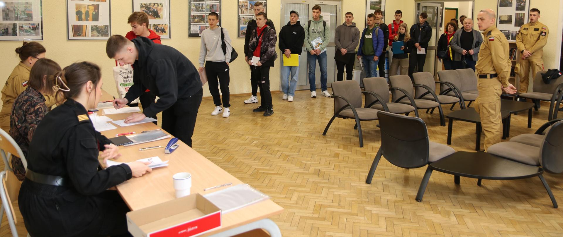 Korytarz (tzw. przejściówka) w budynku Szkoły Aspirantów PSP w Krakowie. Przy stolikach siedzą trzy kobiety, dwie w mundurach, jedna w ubraniu cywilnym. Punkt informacyjny i przyjmowania podań do Szkoły i stojąca do niego kolejka. 