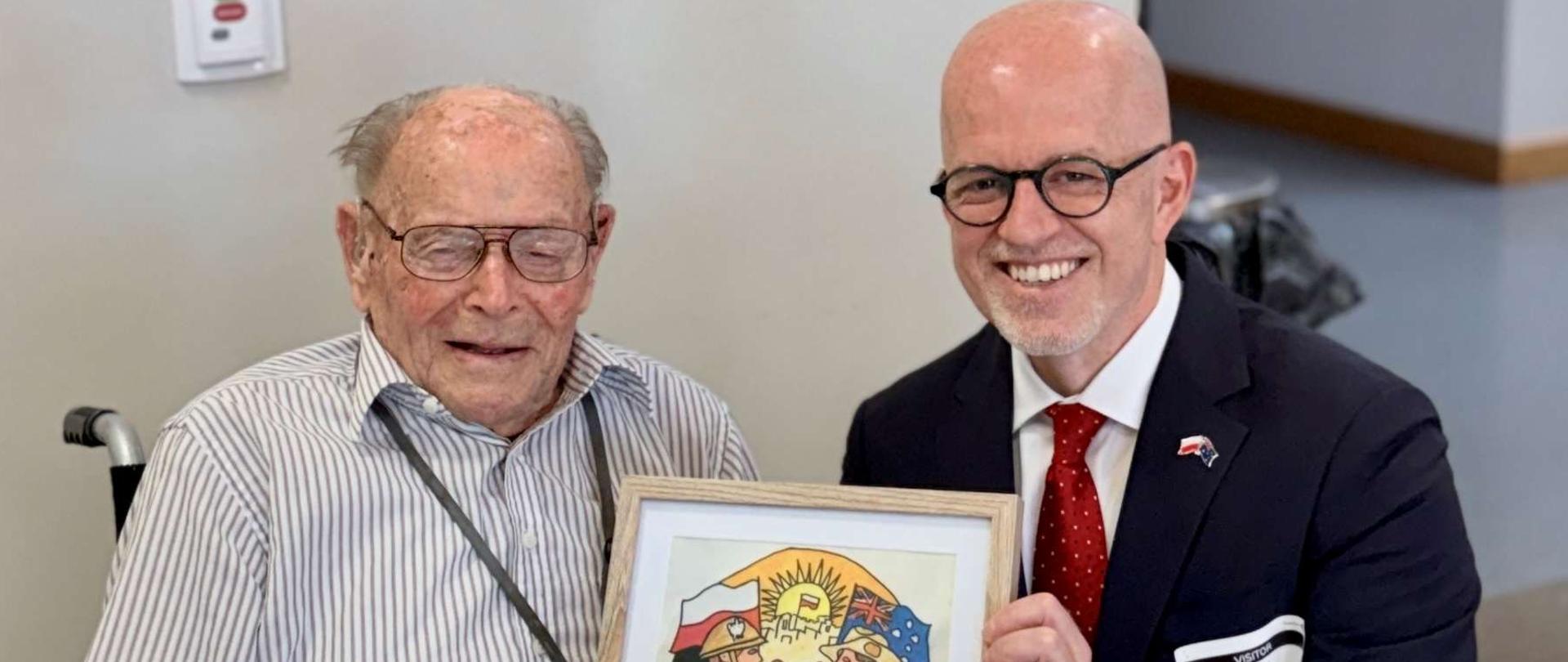 
Two men; the older one is sitting in a wheelchair, while the other,
the younger one, is holding a framed poster
in his hand.
