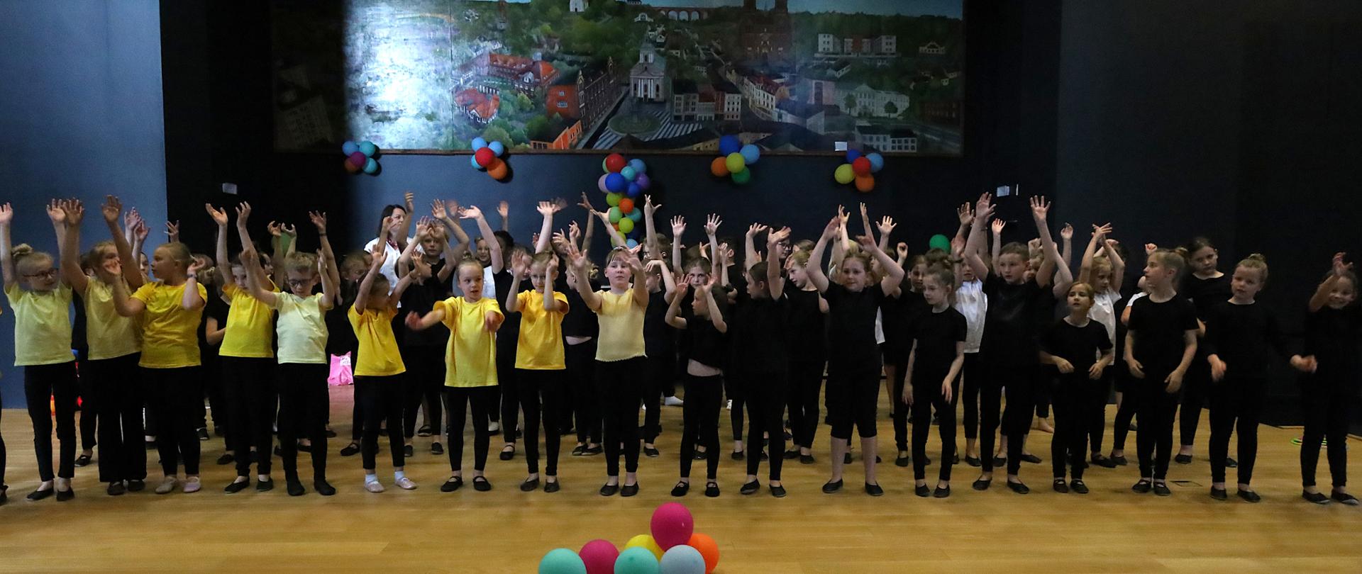Kolorowe zdjęcie na którym znajduję się grupa dzieci ubrana w czarne leginsy i baletki oraz koszulki (żółte, czarne i białe). Dzieci stoją i machają rękami nad głową. Wszystko odbywa się na tle czarnej ściany z dużym, kolorowym obrazem oraz z kolorowymi balonami.